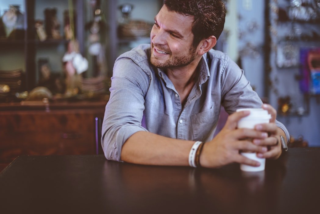 Homem sorrindo segurando copo de café, sentado em mesa de restaurante