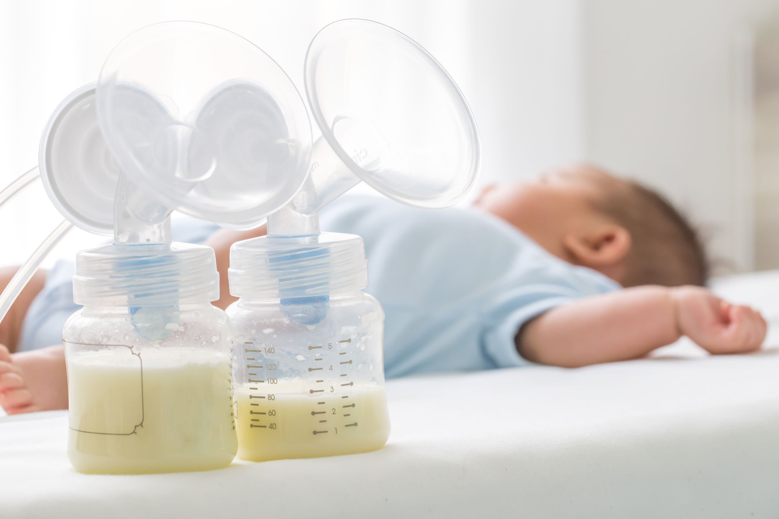 Bebê deitado em cama branca, ao lado há o recipiente de duas bombas elétricas de leite materno