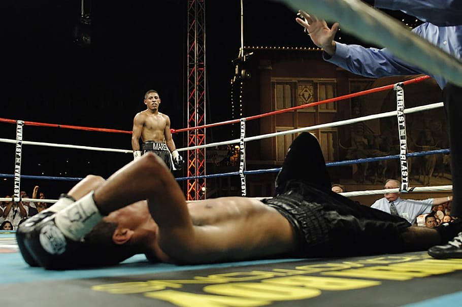 Homem deitado no chão em ringue de luta, com as mãos, cobertas por luvas, em sua cabeça. Ao fundo, outro homem, de pé, também dentro do ringue, observa a situação.