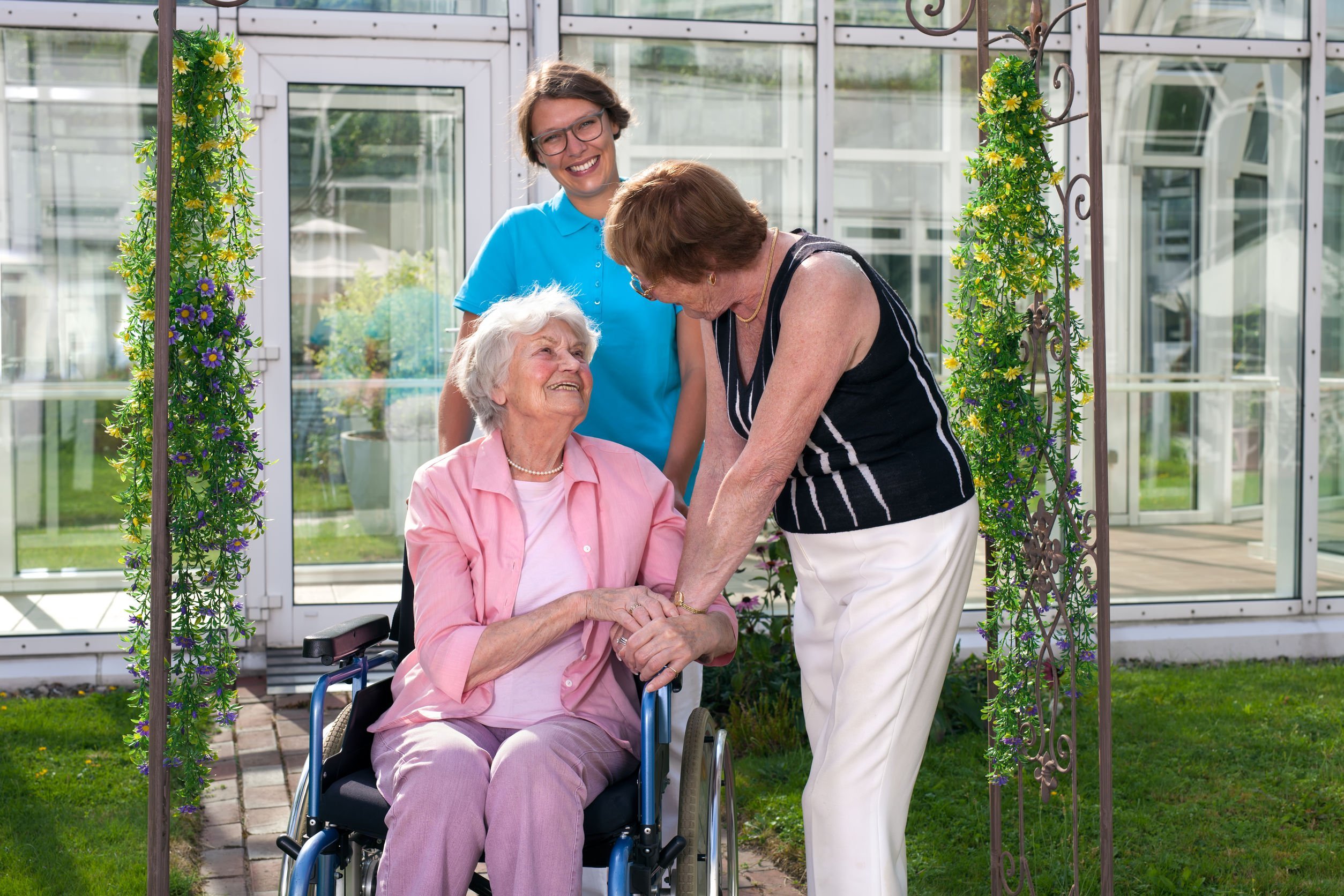 Senhora sentada em cadeira de rodas enquanto uma enfermeira a empurra e uma mulher conversa com ela, as três estão sorrindo