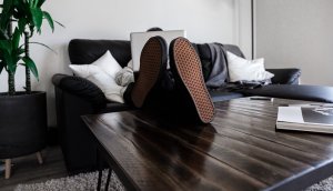 Dillon Shook/ Unsplash Homem sentado em sofá segurando notebook com os pés em cima de mesa de centro