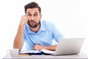 piksel/123RF Homem apoiando cabeça em braço, com expressão de tédio, sentado em mesa com computador, papéis e xícara