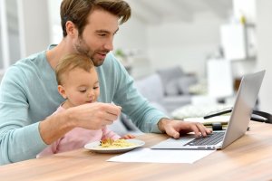 goodluz /123RF Homem dando comida para bebê que esta no seu colo ao mesmo tempo em que mexe em notebook