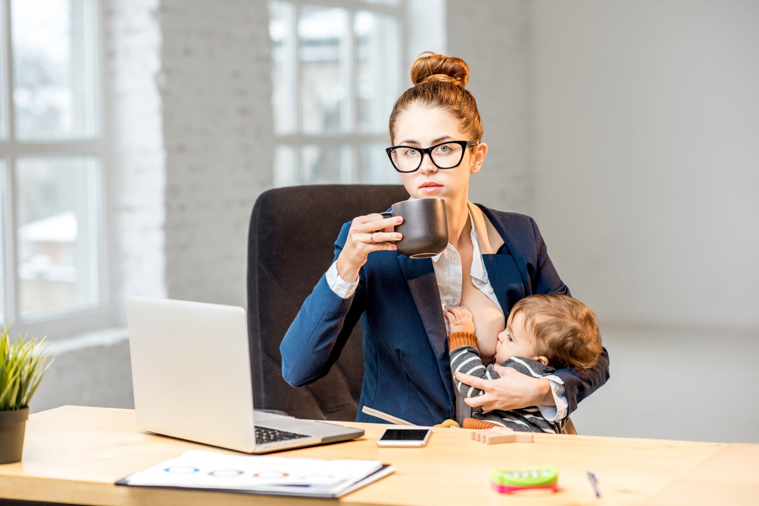 Mulher sentada em mesa de escritório, olhando para notebook, tomando café e amamentando filho