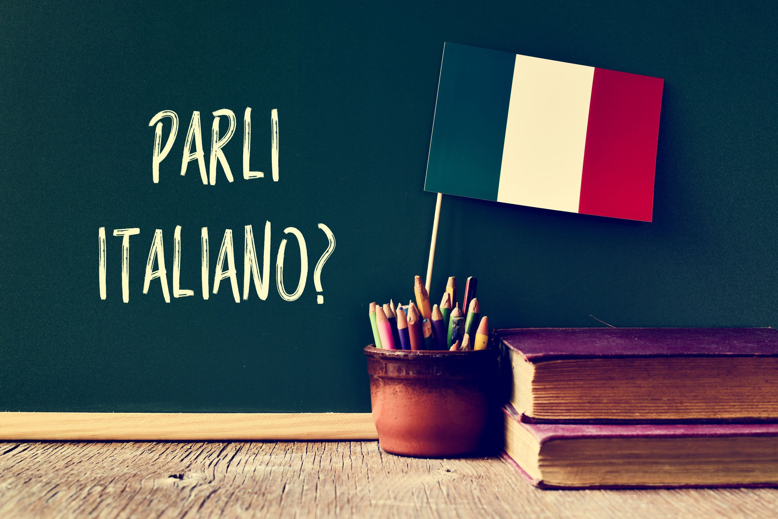 Lousa com a frase 'fala italiano' escrito em italiano atrás de porta lápis com miniatura de bandeira da itália