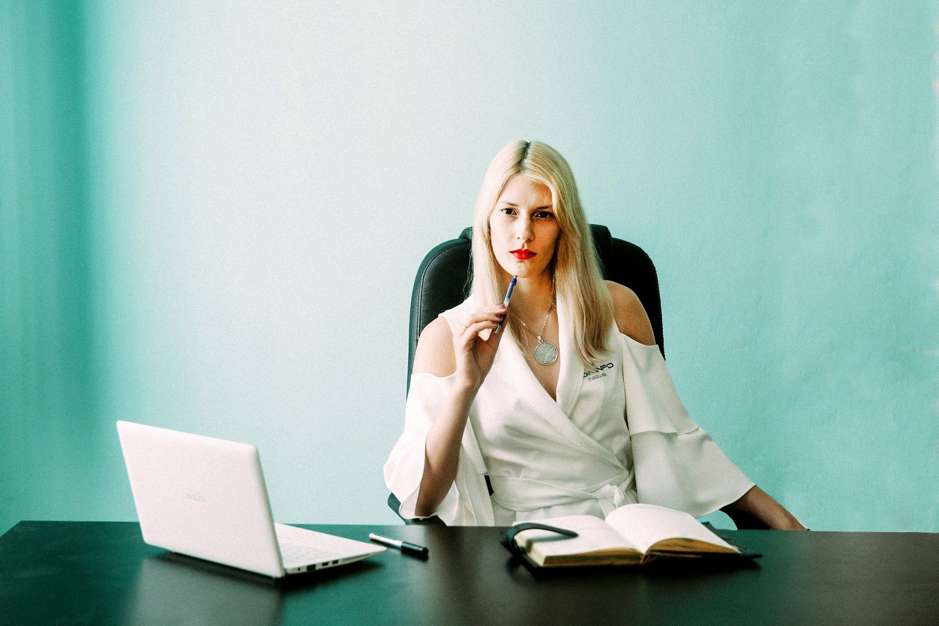 Mulher sentada em mesa de escritório, em frente à notebook, segurando caneta com expressão de dúvida