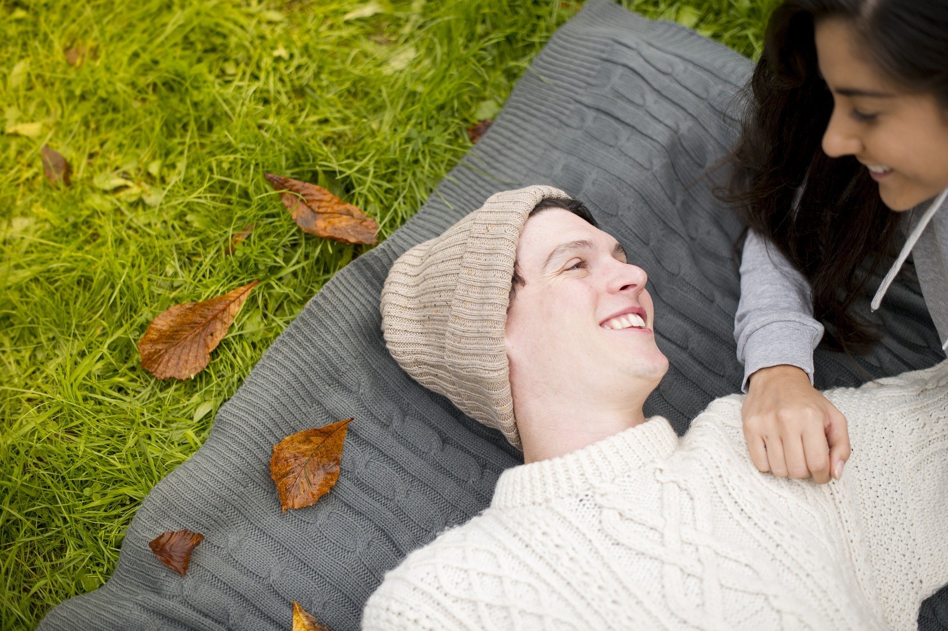 Homem deitado em cobertor sobre gramado, sorrindo, com mulher olhando para ele sorrindo