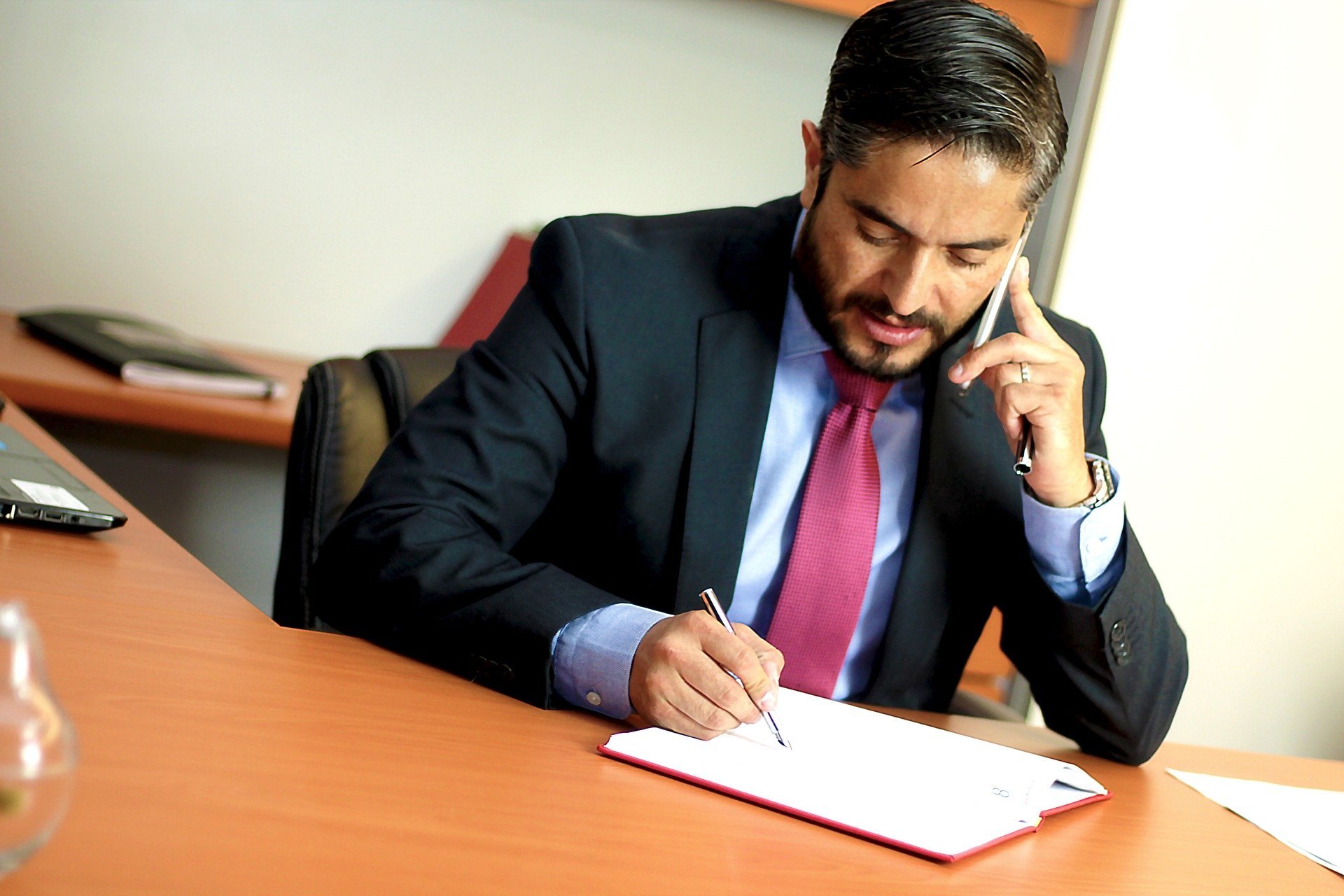 Homem usando roupas sociais, sentado em mesa de escritório, falando no celular e fazendo anotações