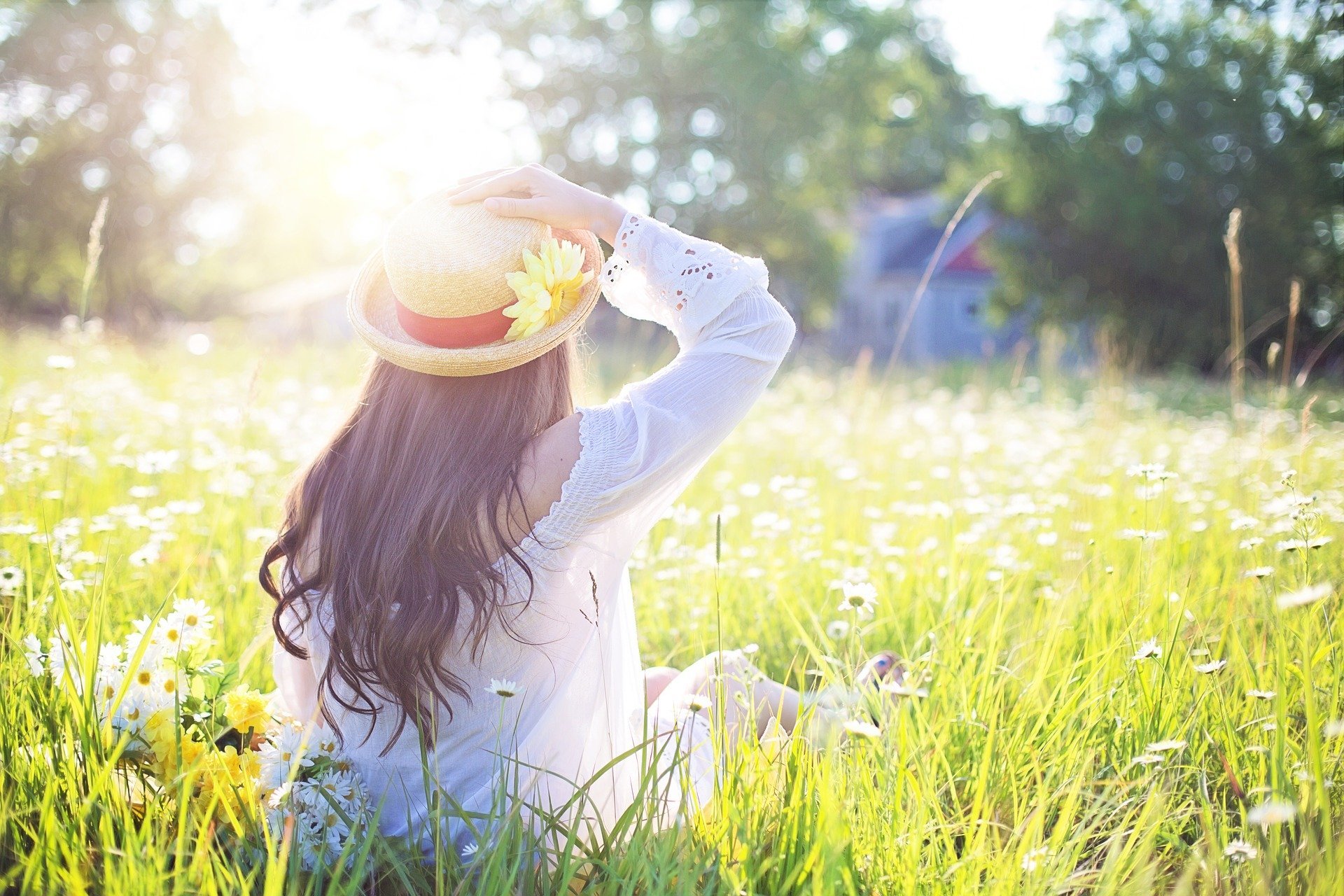 Mulher  sentada em meio de campo de flores com mão segurando chapéu que está em sua cabeça