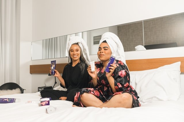 Duas mulheres em uma cama com toalhas na cabeça, segurando produtos de beleza