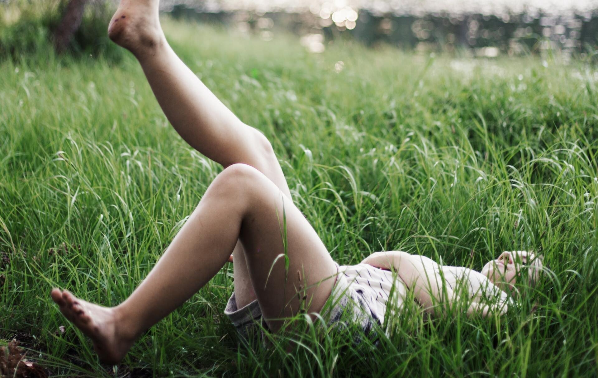 Menina branca deitada na grama alta, com uma de suas pernas levantadas.