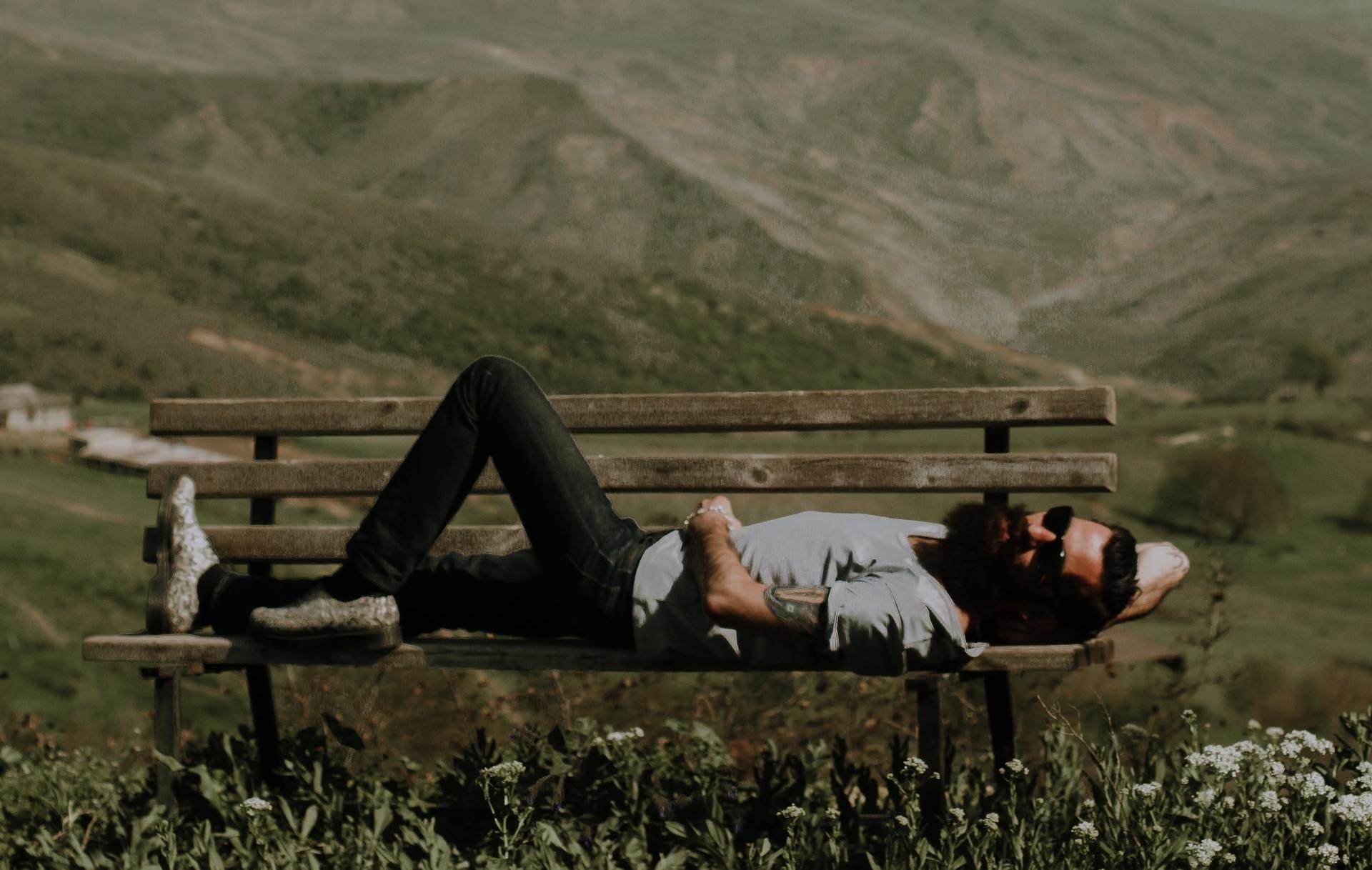 Homem branco de barbas negras, utilizando óculos de sol, deitado num banco, perna esquerda dobrada e braço direito atrás da cabeça