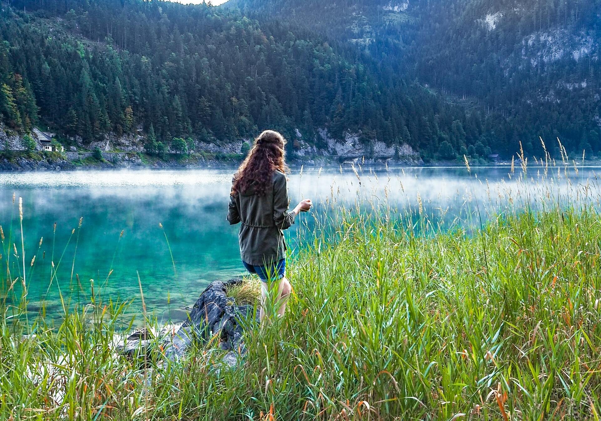 Mulher de cabelos médios e ondulados, de costas em meio a grama, lago e montanhas.