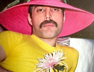 Freddie Mercury de camiseta amarela segurando uma flor e com um grande chápeu de palha