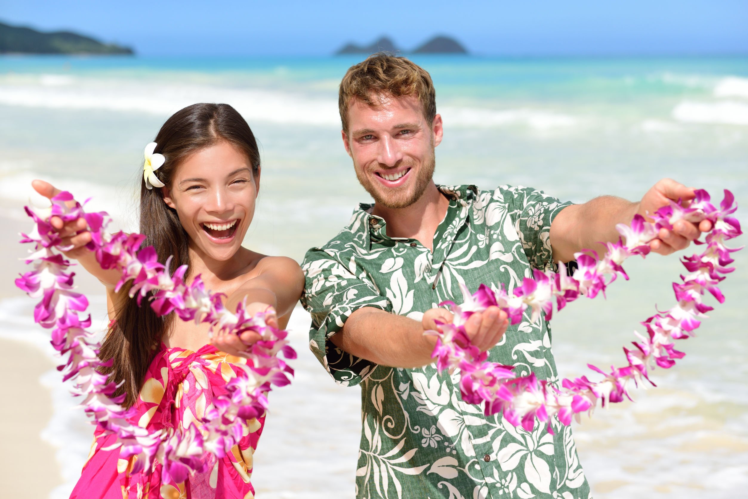Homem e mulher sorrindo, segurando colares havaianos. Em praia.