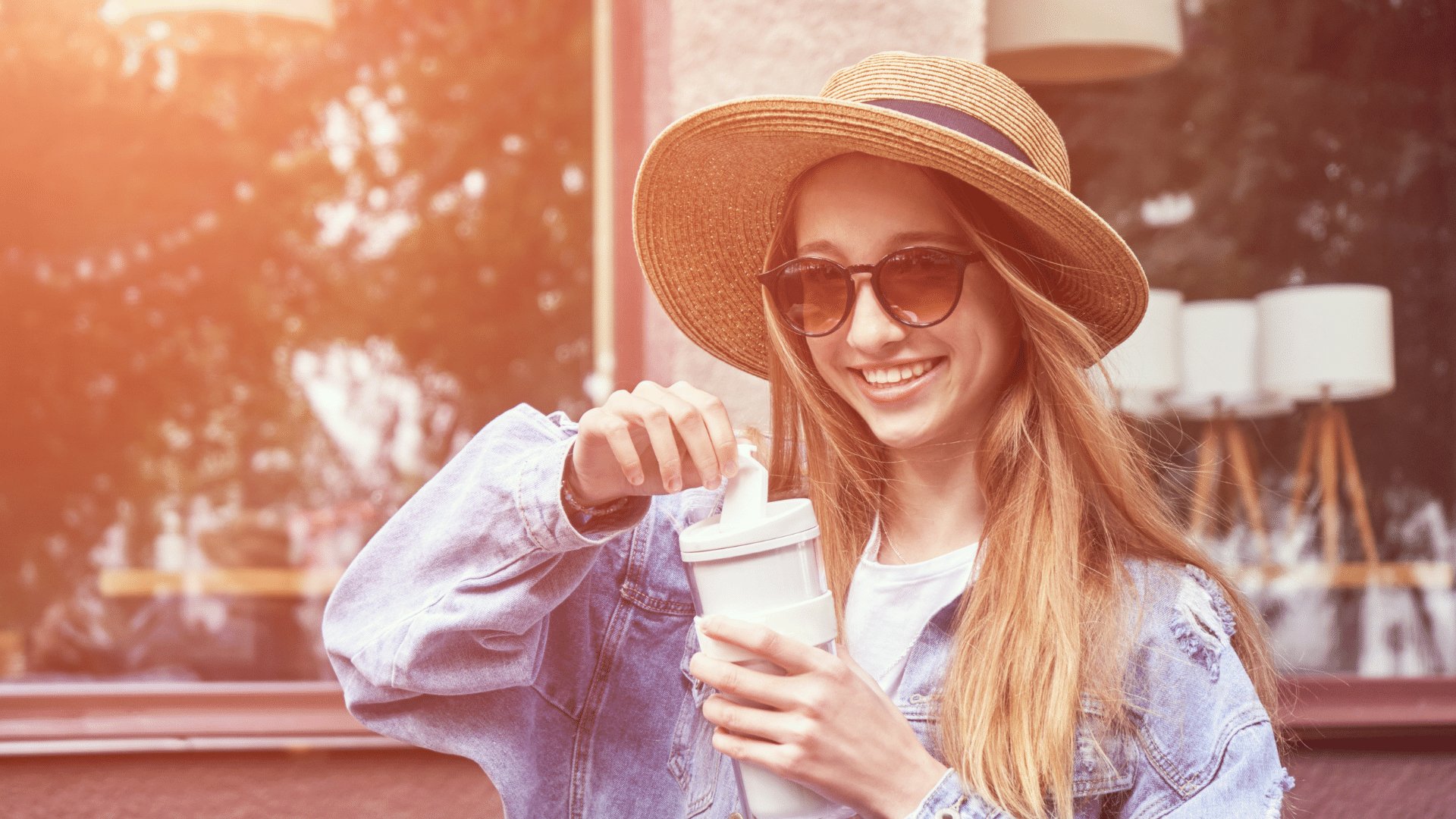 Mulher de óculos de sol e chapéu sorrindo enquanto abre seu copo de café