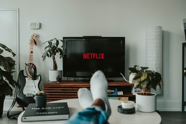 Pessoa com pernas em cima de mesa de canto e tela inicial da Netflix na TV a sua frente