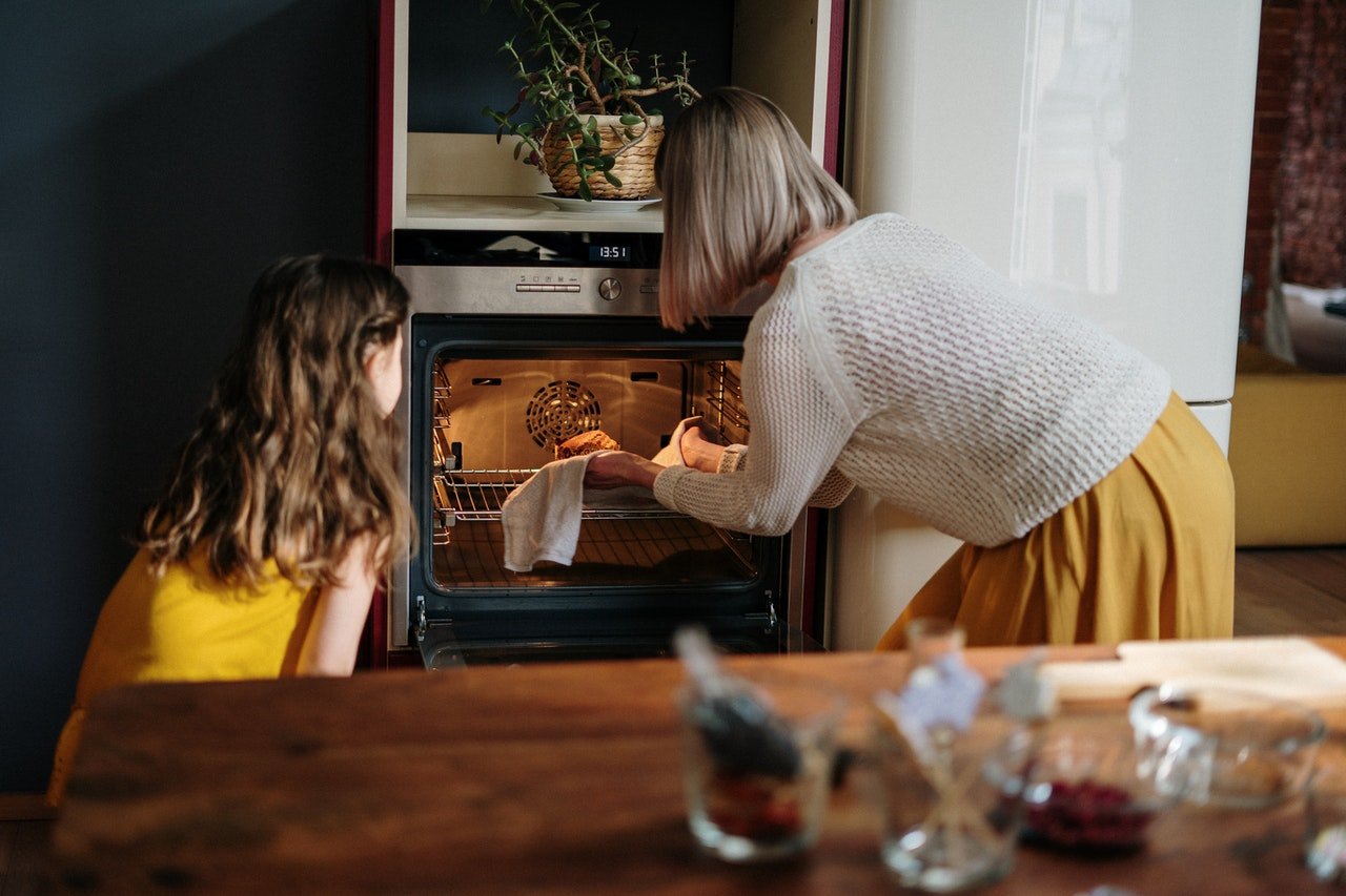 Mãe e filha brancas de costas colocando travessa num forno.
