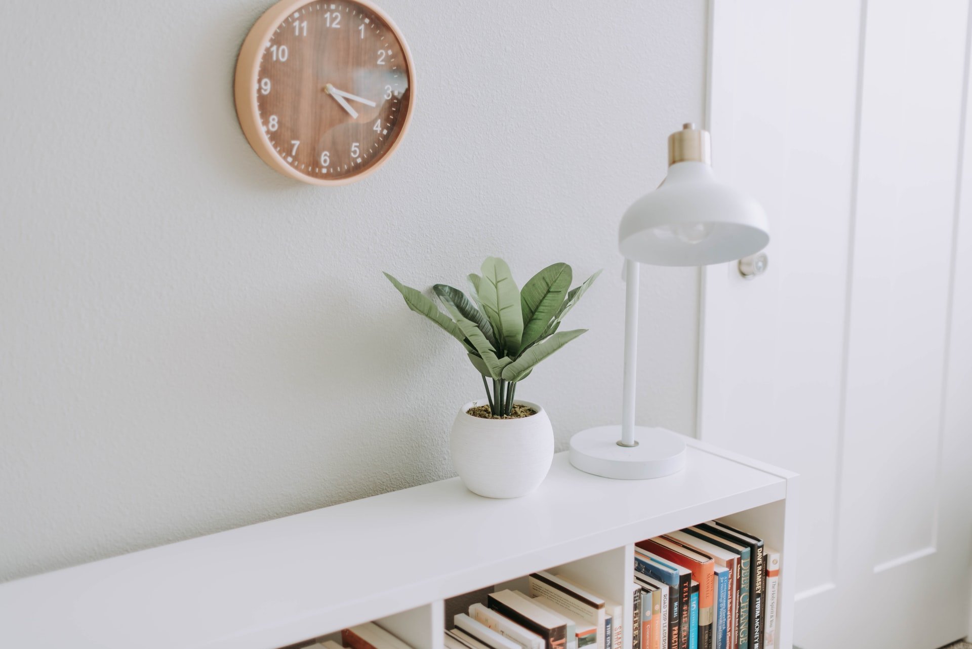 Armário branco com livros, vaso com planta e luminária em cima e um relógio marrom na parede.