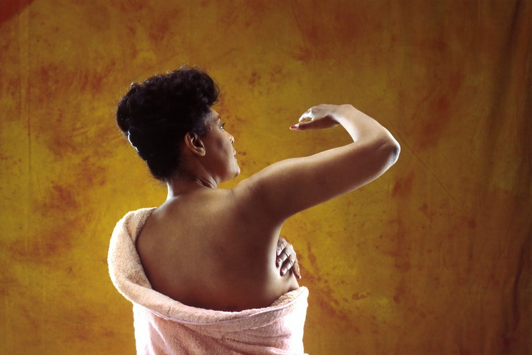 Mulher afro-americana realizando um autoexame das mamas e está vestida com uma toalha a frente um fundo marrom-dourado.