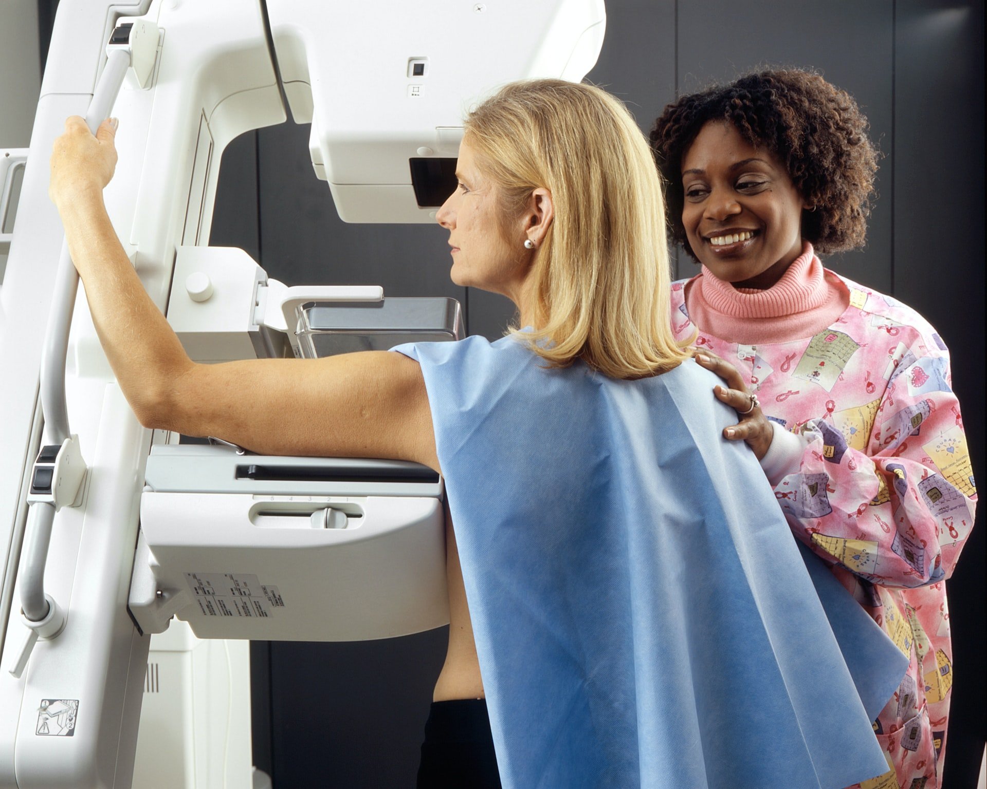 Mulher Recebe Mamografia. Uma técnica afro-americana posiciona uma mulher caucasiana em uma máquina de imagem para receber uma mamografia.