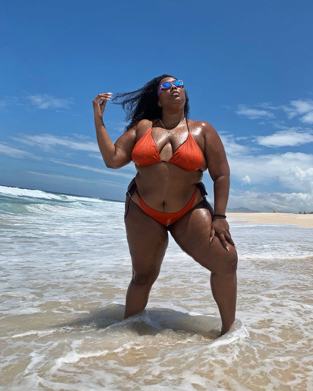 Lizzo na praia com pés na água em pé usando biquini laranja e céu azul ao fundo