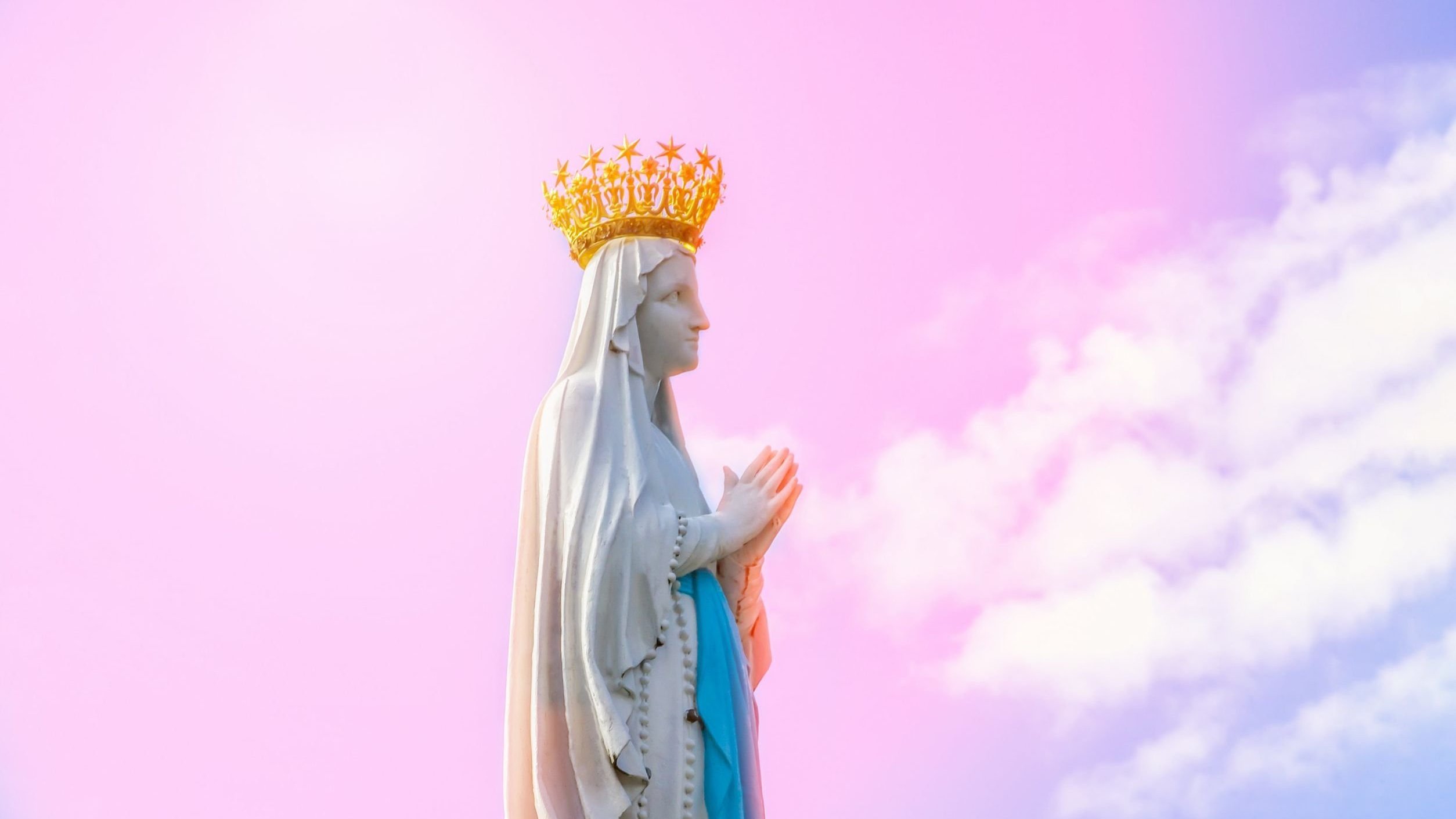 Estátua de virgem Maria de mão unidas, usando coroa. Ao fundo há um céu azul com tons de rosa.