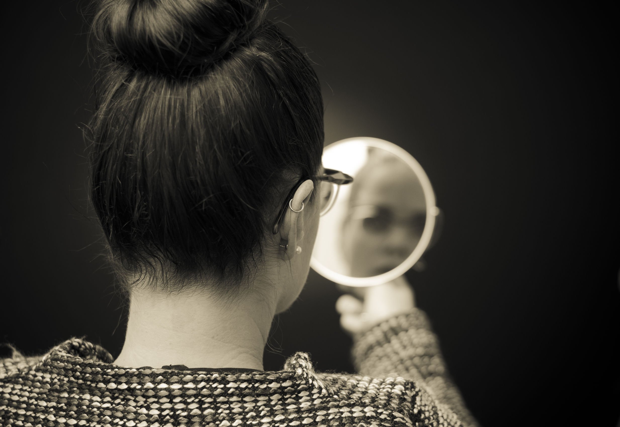 Foto preta e branca de mulher de costas, com os cabelos presos, se olhando em um espelho de mão