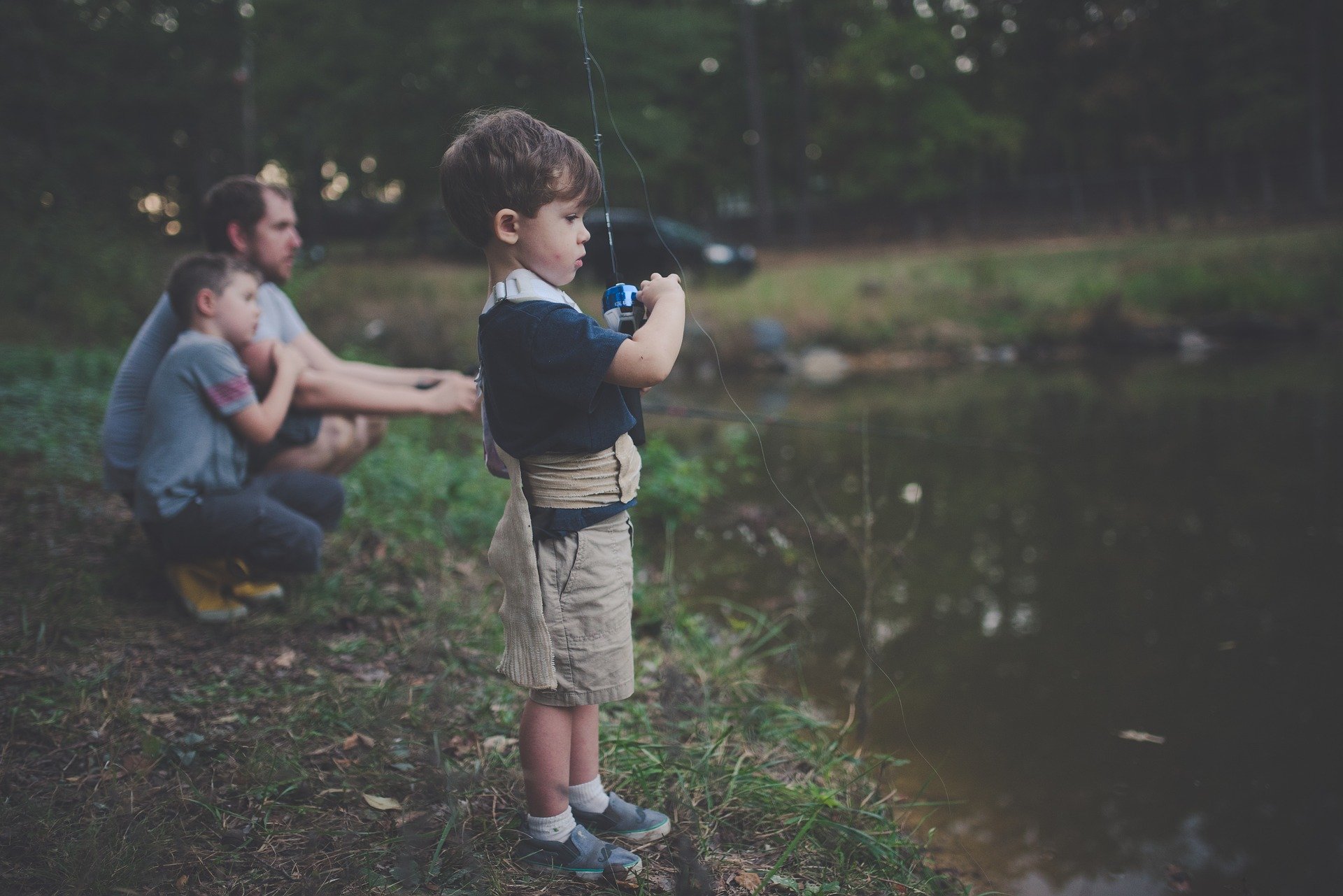 Menino pescando em lago, junto dos pais ao lado.
