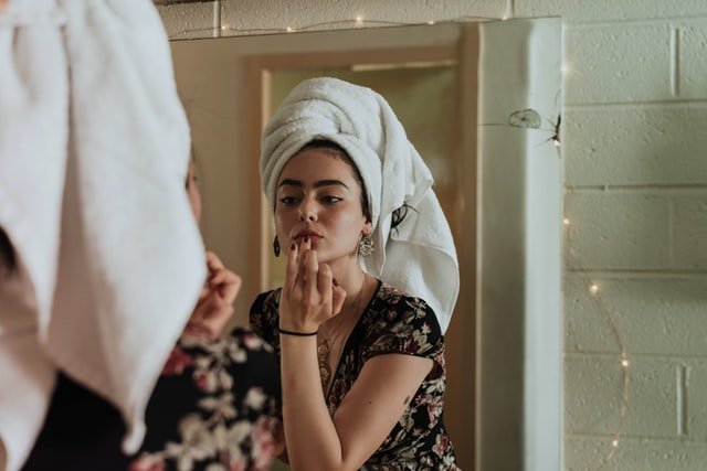 Mulher branca em frente ao espelho com toalha na cabeça e passando batom.