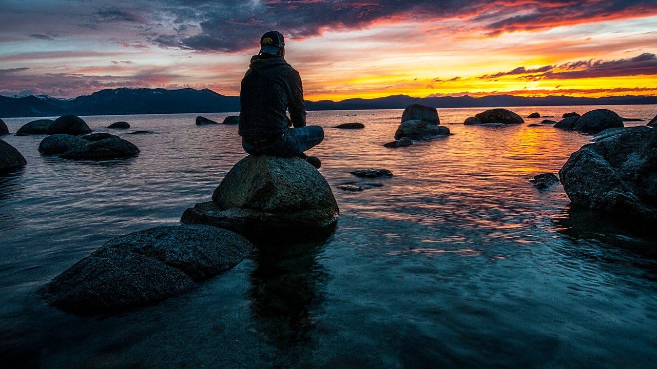 Homem sentado sobre pedra na água a olhar o pôr do sol.