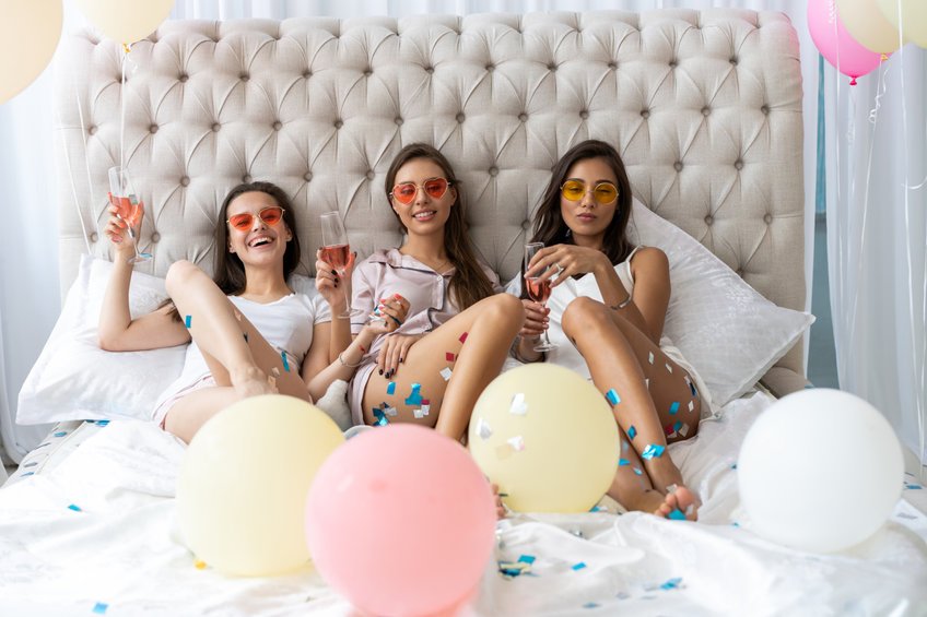 Três mulheres deitadas na cama rodeadas de balões e usando óculos coloridos