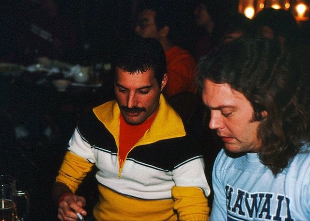 Freddie Mercury de camisa listrada olhando para baixo ao lado de um homem ruivo e cabelo de camiseta cinza
