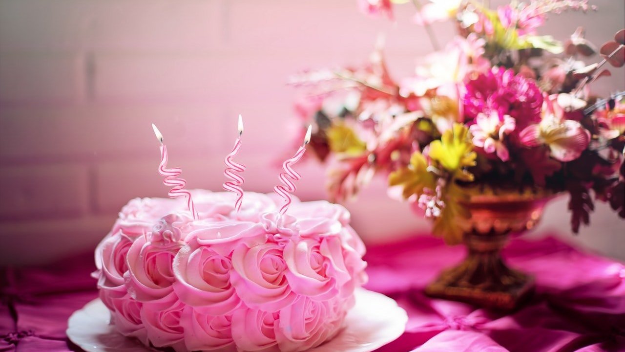 Um bolo cor de rosa com flores no fundo em cima de uma mesa com toalha cor de rosa.