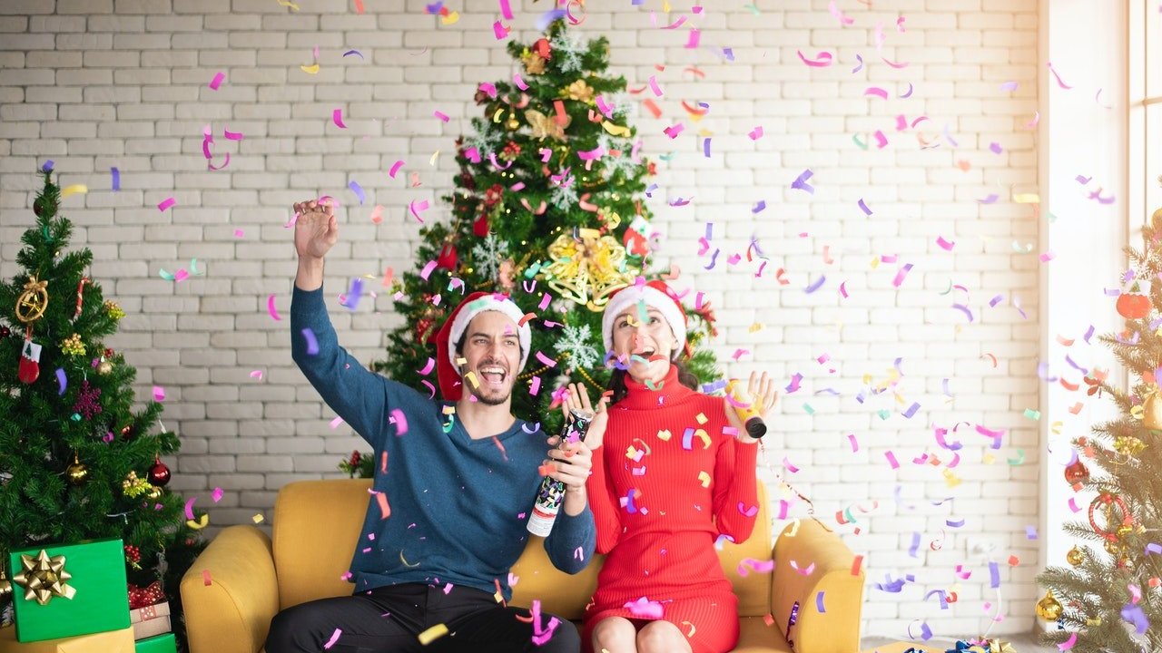 Homem e mulher sentados em sofá comemoram o Natal. Ao fundo, há árvores decoradas e presentes.