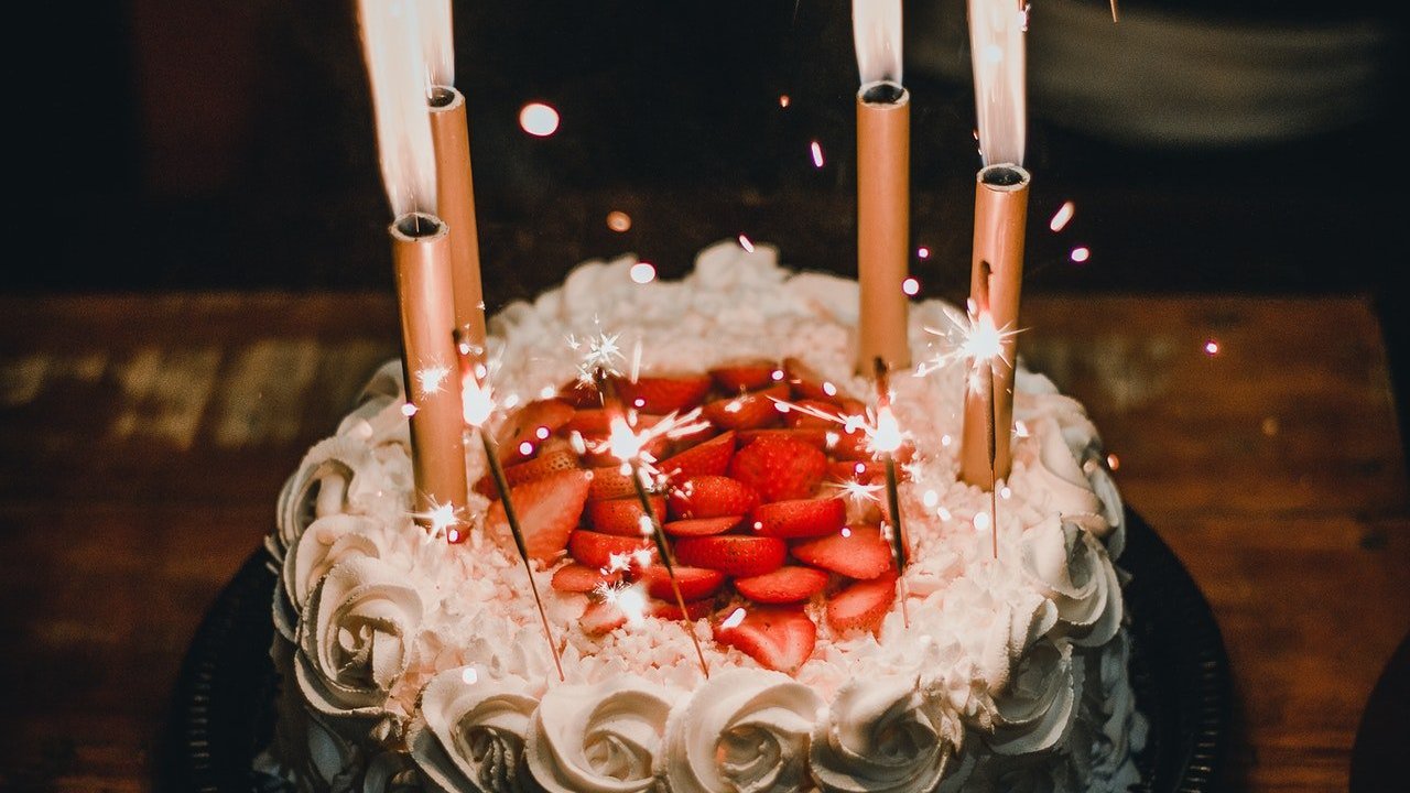Sobre mesa de madeira, bolo redondo decorado com rosas de creme chantili e morangos. Também há velas estrela.