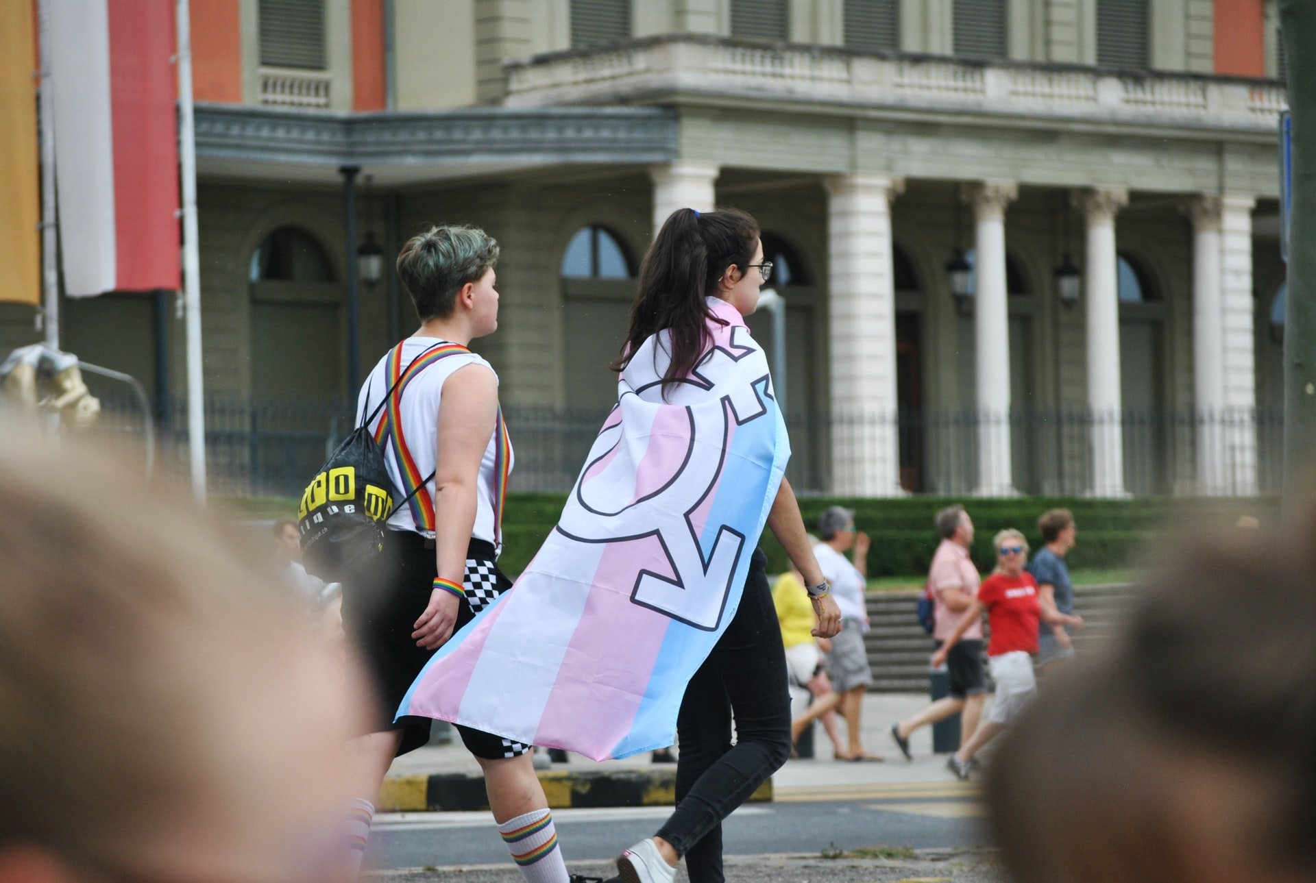 duas pessoas caminhando lado a lado, uma delas com a bandeira trans amarrada nos ombros