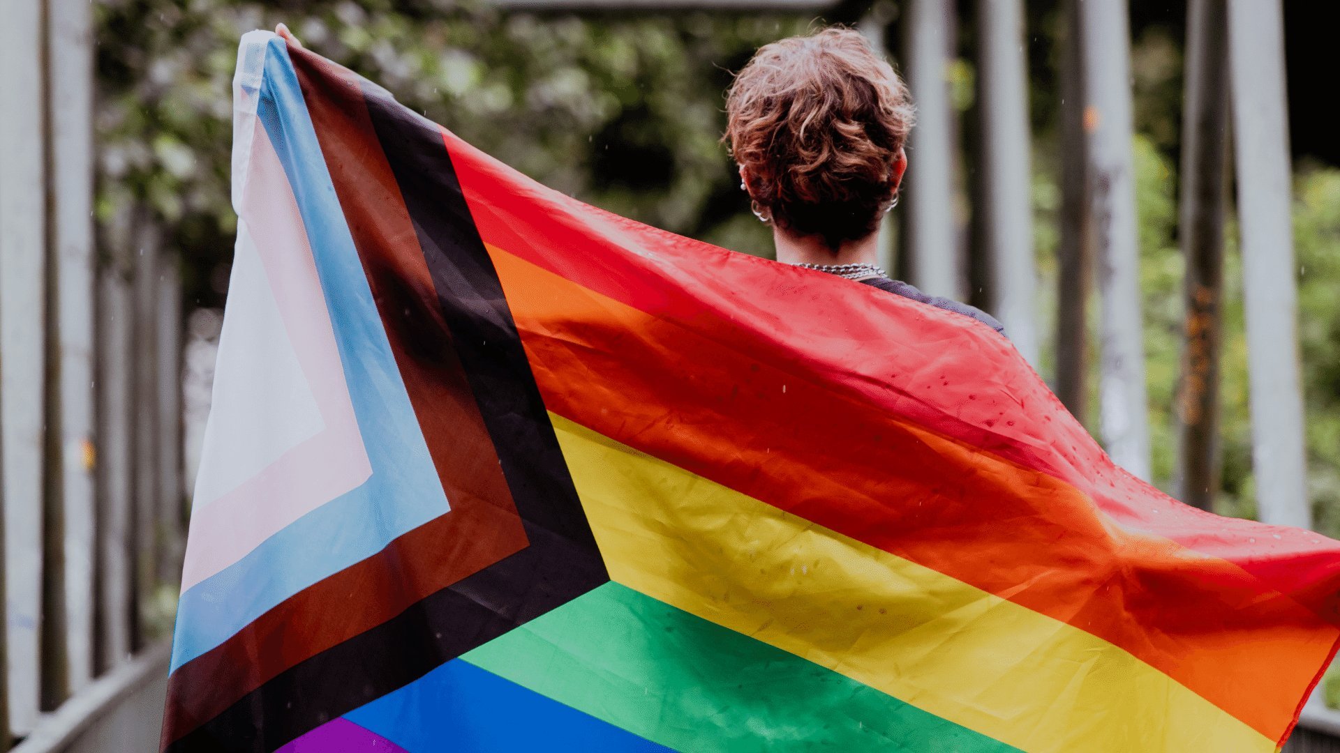 Pessoa de costas segurando bandeira LGBTQI+ com as mãos