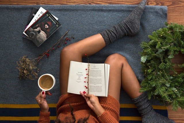 Mulher negra sentada no chão lendo um livro e segurando xícara de café.