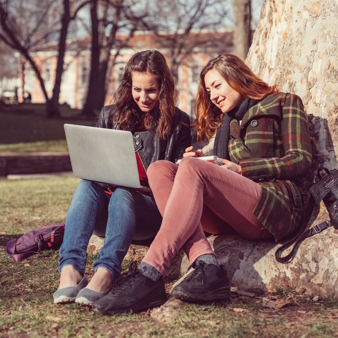Duas mulheres sentadas em tronco de uma árvore, cenário de campus universitário. Uma delas está com um notebook sobre o colo e observa a tela do dispositivo, a outra, ao lado, faz o mesmo.
