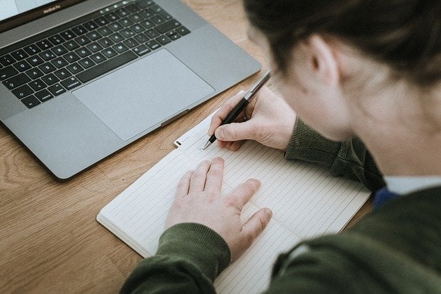 Mulher escrevendo em caderno e notebook em sua frente