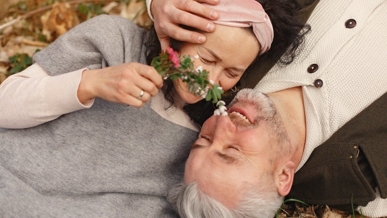 Casal de idosos sorri e se abraça. Eles estão deitados sobre um chão com folhas. A mulher segura um ramo florido próximo do rosto do parceiro, que acaricia a cabeça dela.