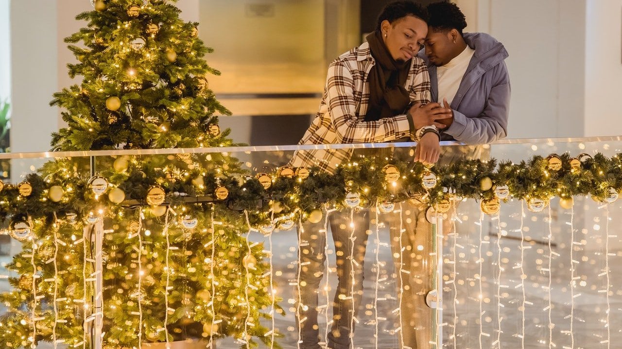 Casal abraçado apoia-se em grade de proteção em sacada decorada com enfeites natalinos. Ao lado deles, há uma árvore de natal.