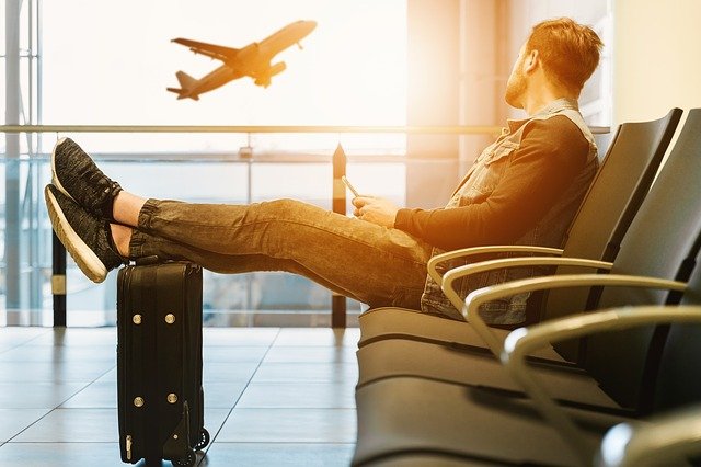Homem com pés apoiado em mala e olhando para avião voando