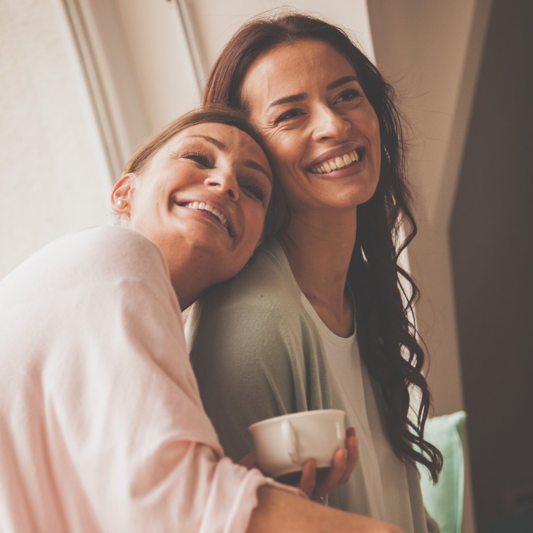 Duas mulheres juntas e sorrindo, enquanto a mulher da esquerda está com a cabeça apoiada no ombro da mulher na direita