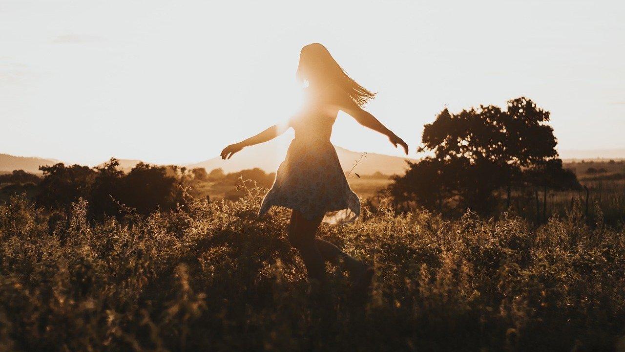 Uma mulher de braços ligeiramente abertos enquanto o sol lhe faz um movimento de contraluz, ofuscando-a do campo de visão de quem a observa. Ao fundo, árvores e um gramado fértil.