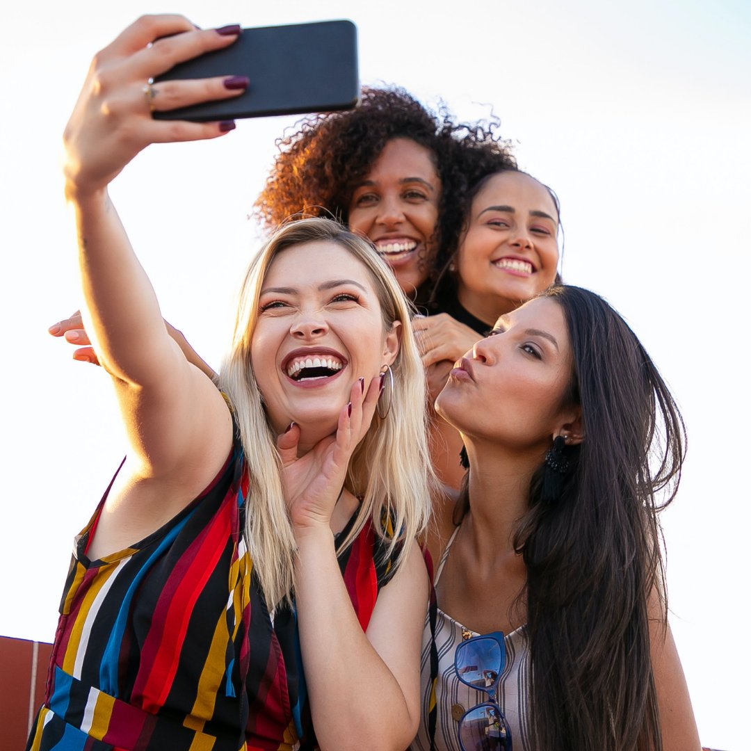 Uma mulher loira tirando uma selfie com outras pessoas ao fundo: uma mulher negra ao fundo, e outros duas mulheres morenas à direita.