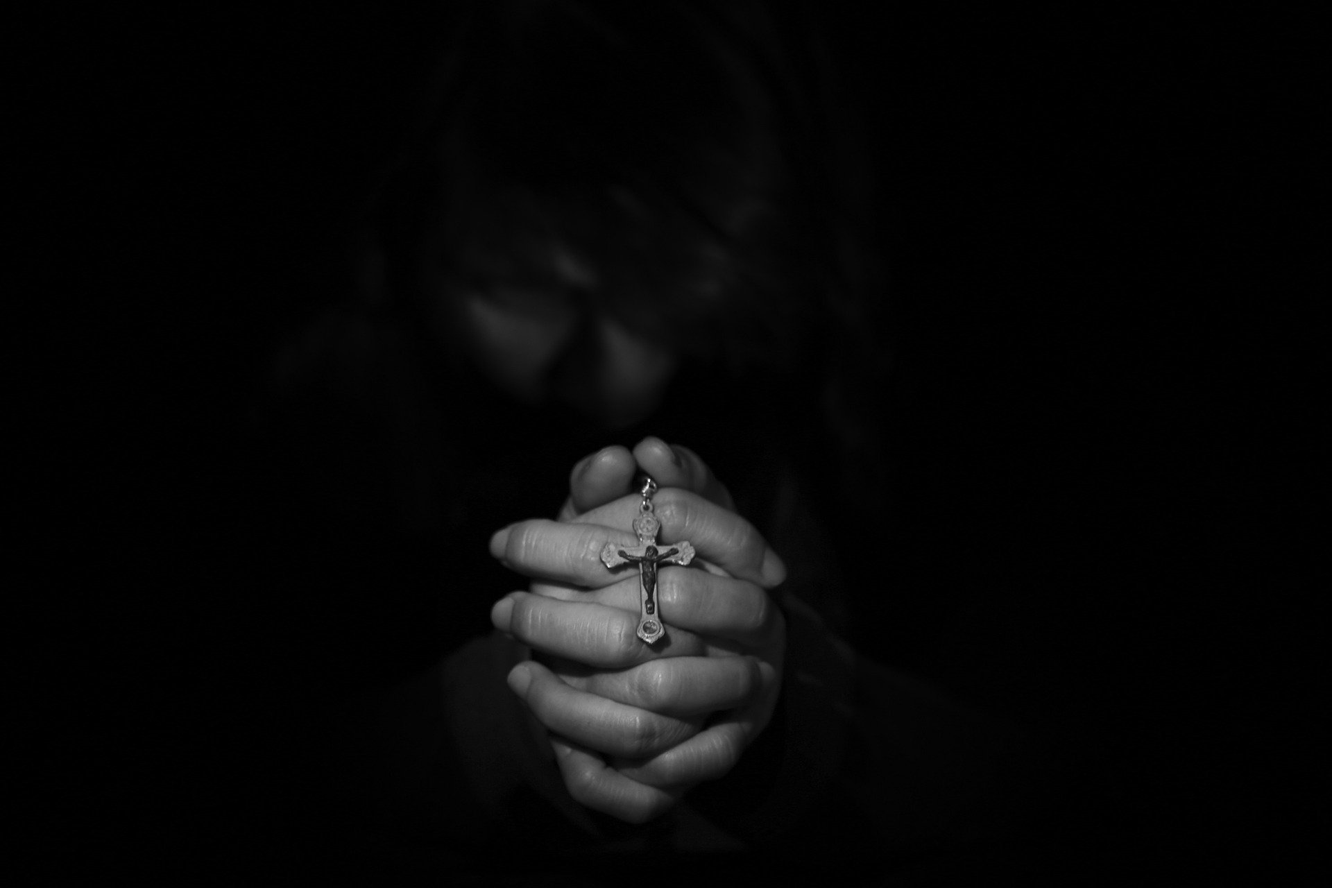 Uma pessoa curvada, em gesto de oração, no escuro. Nas suas mãos, um pequeno terço cristão.