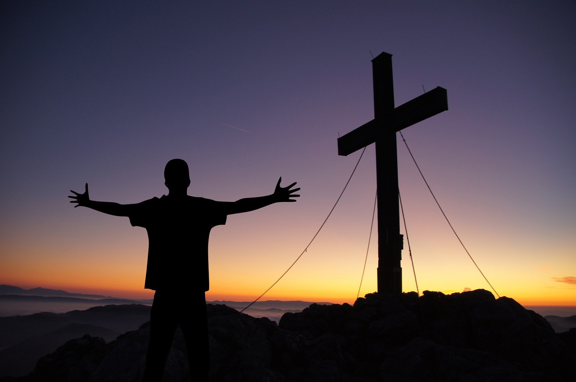 Uma silhueta masculina de braços abertos contemplando uma cruz no pôr-do-sol.