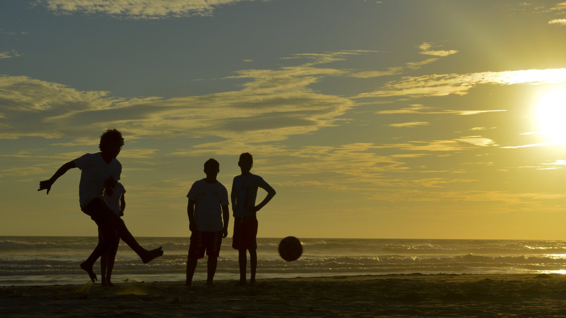 Pessoas jogando futebol numa praia que exibe um por do sol.